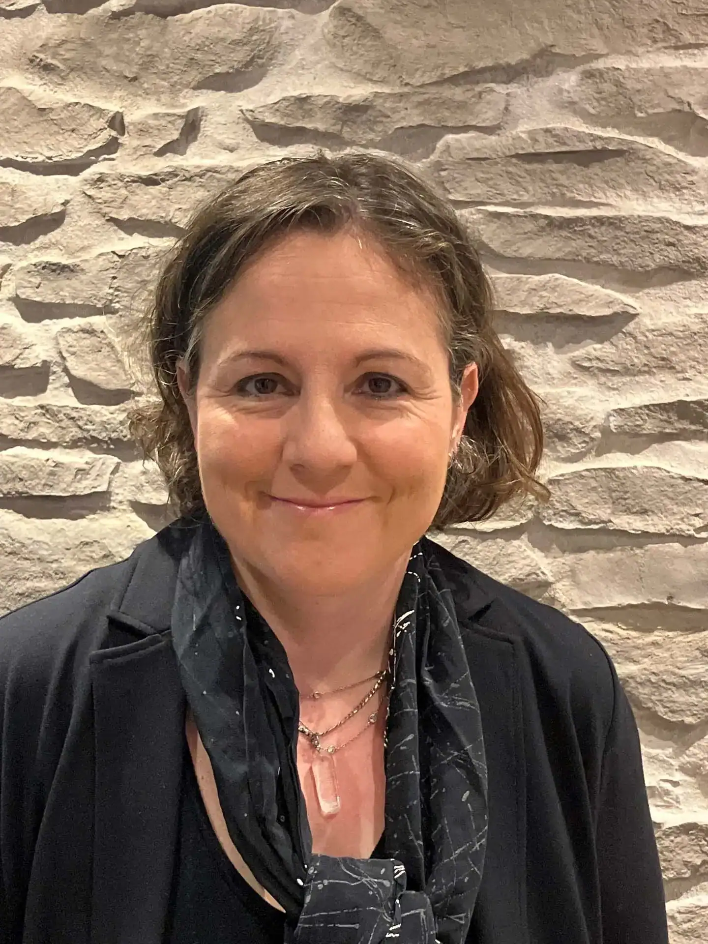 Eine lächelnde Frau mit kurzen braunen Haaren steht vor einer strukturierten Steinwand. Eine lächelnde Frau mit kurzen braunen Haaren steht vor einer strukturierten Steinwand.